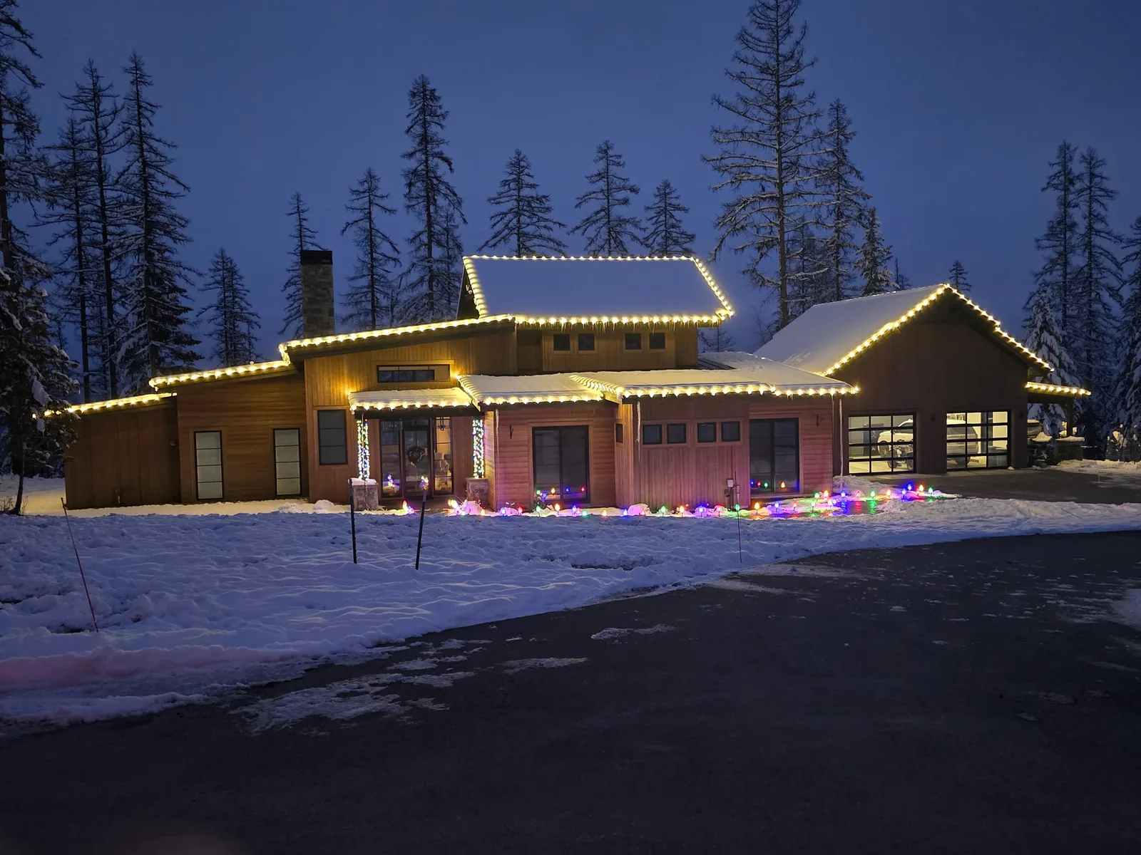 A Flathead Valley home with warm-white roofline lighting photographed at blue-hour dusk, tall pines behind the house